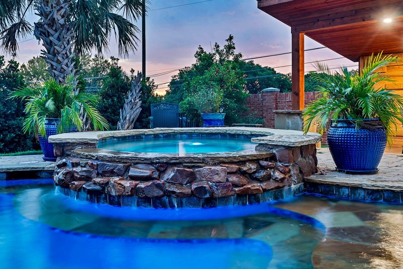 Lit spa spilling over a stone wall into the pool at dusk